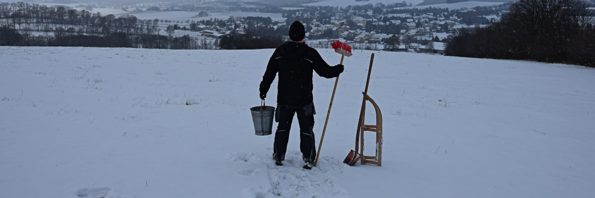 Marek, der Wintermacher – Mann mit Schlitten, Besen und Eimer steht auf verschneitem Feld und blickt prüfend auf Großwaltersdorf, als würde er den Winter ausliefern