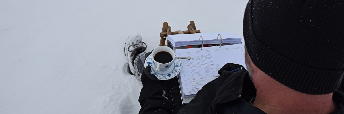 Technikpause im Schnee – Mann sitzt auf einem Schlitten mit Kaffee und studiert einen Elektroplan im verschneiten Erzgebirge
