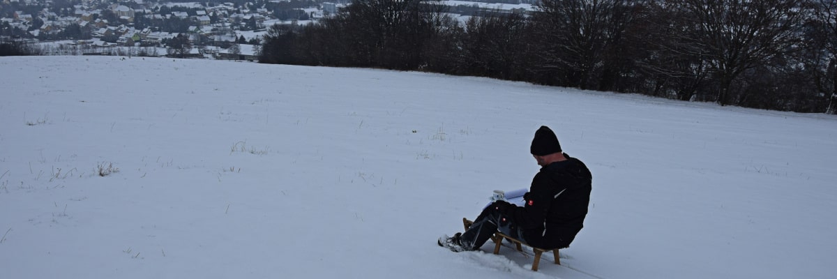Technikpause im Schnee – Mann mit Schlitten und Kaffee studiert einen Elektroplan in verschneiter Landschaft